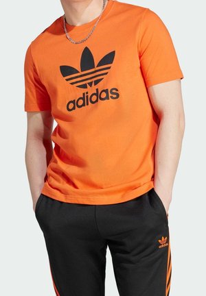 Oranje katoenen T-shirt met een zwart Adidas-logo en drie zwarte strepen in een bladachtig ontwerp, gecombineerd met zwarte broek en oranje accenten.