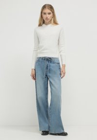 Frau steht in einem weißen Strickpullover, hellblauen weit geschnittenen Jeans mit gebundenem Bund und schwarzen Schuhen vor einem schlichten weißen Hintergrund.