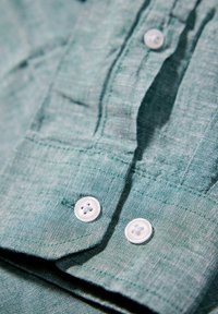 Ljusgrön linne skjorta med en texturerad väv. Har två vita knappar framtill och vikta manschetter, vilket framhäver tygets mjukhet.