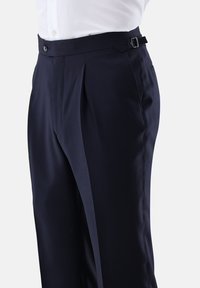 Marineblaue Anzughose mit seitlicher Verstellschnalle, vorderen Falten, Knopfverschluss, getragen mit einem in die Hose gesteckten weißen Hemd.