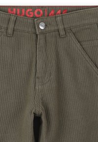 Olijfgroene ribbroek met een rood merklabel en een zilveren knoop bij de taille; voorzien van zakken met versterkte stiksels.