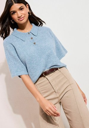 Fiatal nő világoskék rövid ujjú gombos pulóverben és bézs nadrágban, barna bőr övvel, világos fal előtt áll.