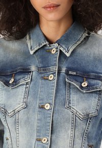 Chaqueta de denim azul claro con cuello extendido, dos bolsillos en el pecho y botones en tono plateado. La textura desgastada añade un interés visual.