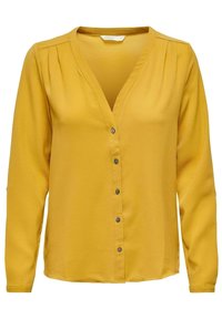 Blouse manches longues jaune moutarde avec col en V, fermeture par boutons, détail de plis pincés aux épaules et tissu texturé.