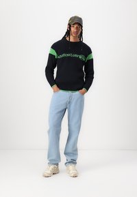Pulover negru cu dungi și accente verzi și logo, blugi de culoare albastru deschis și adidași bej. Modelul poartă o șapcă de camuflaj. Fundal simplu.