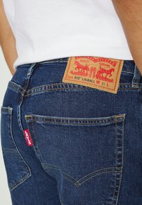 Mörkblå denimjeans har en tan läderpatch med röda grafiker och vit text, samt en liten röd etikett på bakfickan.