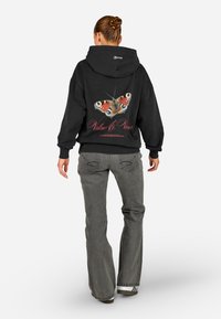 Mulher com um coque, vestindo um hoodie preto com uma grande borboleta colorida e a inscrição vermelha "Valores & Visões" nas costas, jeans cinzentos e sapatilhas.