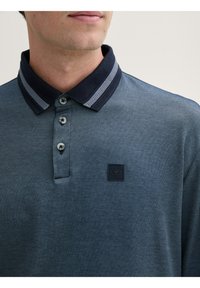 Koszulka polo w ciemnoniebieskim kolorze z fakturowanej tkaniny, z kontrastującym granatowym kołnierzykiem i zapięciem na trzy guziki. Zawiera małą naszywkę z logo.