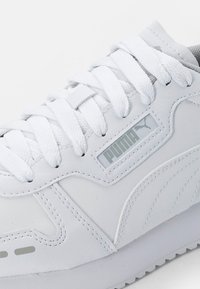 White sneaker with a smooth leather upper, lace closure, grey accents, and a prominent logo. Features textured paneling and a cushioned sole.