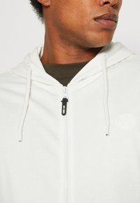 Bjela zip-up dukserica napravljena od mekanog materijala, sa prednjim zipom, kapuljačom s vezicama i malim izvezenim logotipom na grudima.