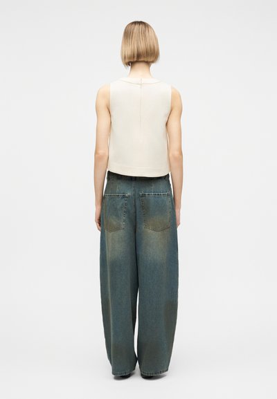 Personne aux cheveux blonds courts portant un haut blanc sans manches et un jean bleu délavé à jambes larges, debout de dos sur un fond uni.