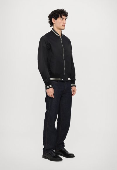 Giubbotto bomber nero con colletto e polsini a costine, chiusura con zip, abbinato a jeans denim scuri e scarpe nere. Texture liscia, design semplice.
