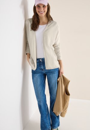 Junge Frau lächelt, trägt eine lila Mütze, eine beige Strickjacke über weißem Oberteil, blaue Jeans, hält eine beige Jacke, steht vor einer weißen Wand.