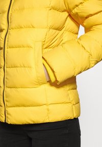 Żółta pikowana kurtka z przednimi kieszeniami, noszona na zamek, z ręką włożoną do prawej kieszeni, w połączeniu z czarnymi spodniami.