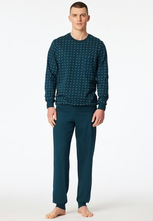 Young man standing barefoot wearing a dark teal long-sleeve patterned sweater and matching solid teal pants against white background.
