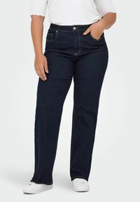 Tumši zili džinsi ar taisnu kāju piegriezumu, kuriem ir priekšējās kabatas un smalki šuvju akcenti. Valkāti ar baltu augšu un baltām kurpēm.