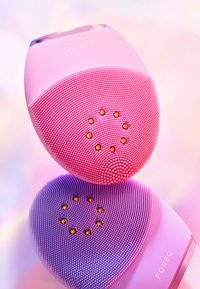 Nettoyant facial en silicone aux teintes roses et violettes, avec des surfaces texturées et de petites brosses, et des accents dorés. Forme ovale compacte.