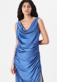 Kumarükkäe sinine satiin kleit, kus on külgedelt rippuvad detailid ja reie kõrge lõhik, pehme tekstuuri ja elegantse siluetiga.