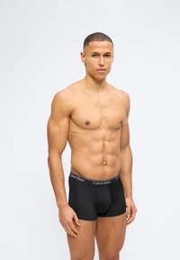 Μυώδης άνδρας με κοντά μαλλιά που φοράει μαύρα μπόξερ της Calvin Klein, στέκεται μπροστά σε απλό ανοιχτό φόντο.