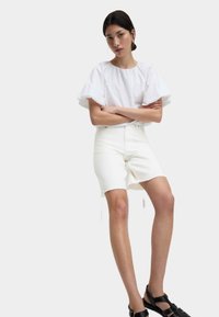Blusa bianca con maniche a palloncino, abbinata a pantaloni corti color crema e sandali neri con cinturini. Lo sfondo è grigio uniforme.