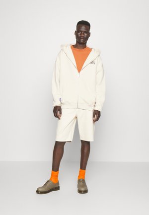 Cremfarbene Zipp-Hoodie mit großer Kapuze, orangefarbenes T-Shirt, weiße Shorts, orangefarbene Socken und beigefarbene Schuhe mit gelben Akzenten.