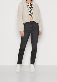 Personne portant une veste courte texturée crème, un crop top à motifs noir et blanc, un jean noir, des chaussettes blanches et des baskets blanches, debout contre un mur blanc.