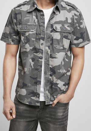 Mand iført kortærmet skjorte med gråt og sort camouflageprint over en hvid t-shirt, med den ene hånd i lommen og grå jeans.
