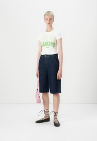 Hvit t-skjorte med grønn grafisk tekst, mørk denim bermudashorts, svarte snøresko, og en rosa håndveske med sølvdetaljer.