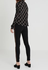 Blusa nera a fantasia con un design a diamante, abbinata a leggings neri con una striscia marrone. L'outfit è completato da tacchi neri.