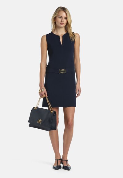 Jeune femme portant une robe sans manches bleu marine avec une ceinture, des chaussures pointues noires, et tenant un sac à main noir avec des chaînes dorées.