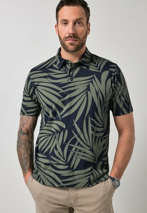 Mężczyzna w ciemnozielonej i czarnej polo w liście z krótkim rękawem, beżowe spodnie, zegarek na lewym nadgarstku i tatuaż na prawym przedramieniu.