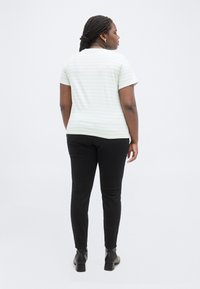 Γυναίκα με κοτσίδα που φοράει ανοιχτόχρωμο ριγέ κοντομάνικο πουκάμισο, μαύρο παντελόνι και μαύρα μποτάκια αστραγάλου, όρθια με την πλάτη στραμμένη.