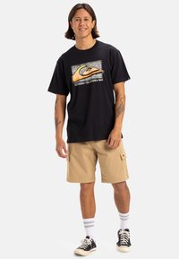 T-shirt nera con stampa grafica bianca e arancione, abbinata a pantaloni cargo beige, calzini bianchi a righe e sneakers nere. Design casual.
