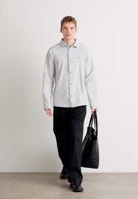 Lys grå skjorte med sorte knapper, båret med svarte bukser. Modellen bærer en stor, svart tote-veske. Enkel og minimalistisk design.