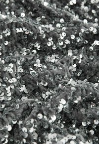 Stříbrné flitry na texturované šedé tkanině, hustě uspořádané v různých velikostech, vytvářející třpytivý a odrazivý povrch.