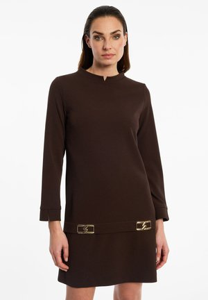 Vrouw draagt een donkerbruine jurk met lange mouwen met gouden gespdetails bij de zoom, staand tegen een effen witte achtergrond.
