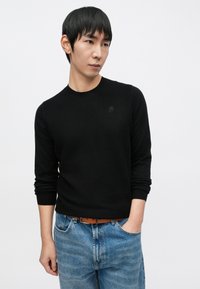 Juodos spalvos megztinis su apvalia iškirpte, ilgomis rankovėmis ir subtiliu logotipu ant krūtinės, derinamas su šviesiai mėlynais džinsais iš džinsinio audinio.