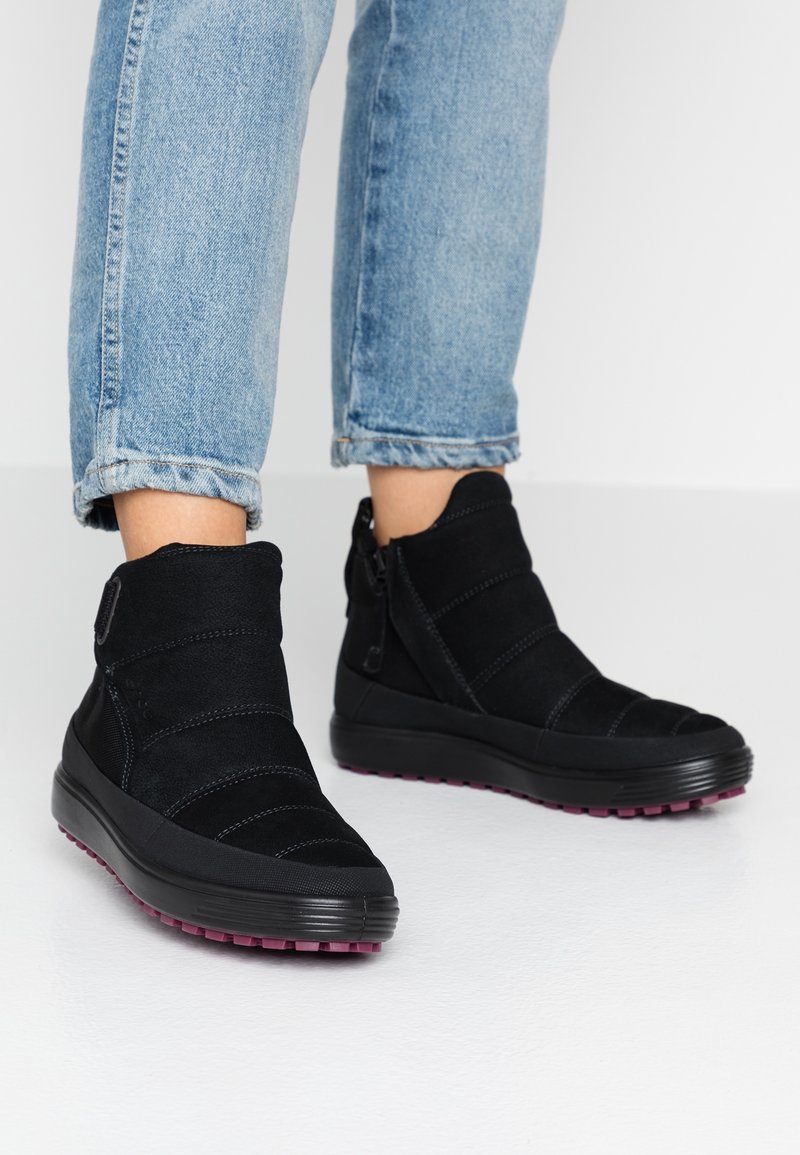 Bottes noires à la cheville avec des zips sur les côtés et semelles violettes, portées avec un jean bleu clair retroussé sur un sol blanc.