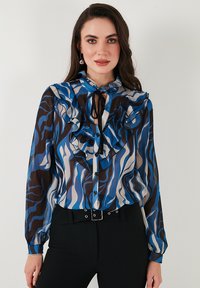 Long-sleeve blouse with ruffled details in blue, brown, and cream wave patterns. Features button-down front and a neck tie.
