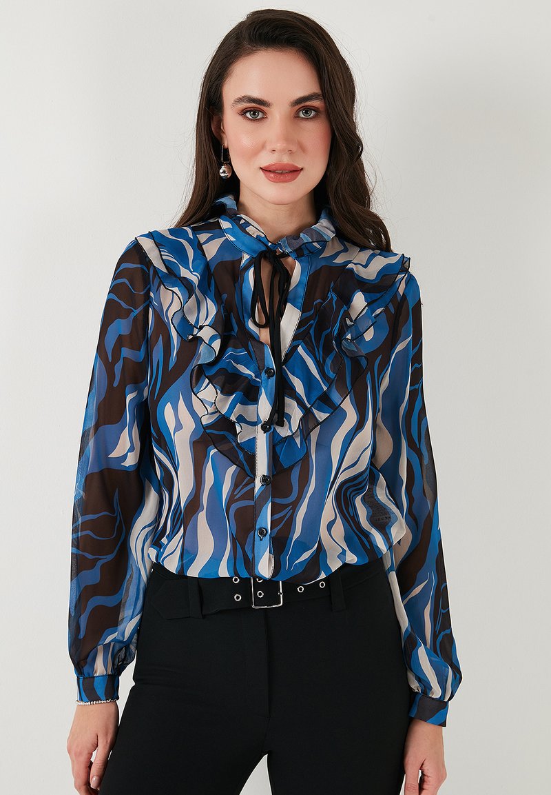 Long-sleeve blouse with ruffled details in blue, brown, and cream wave patterns. Features button-down front and a neck tie.