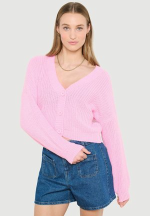 Žena oblečená v svetloružovom pletenom kardigáne na gombíky a modrých riflových krátkych nohaviciach, stojaca s jednou rukou vrecku.