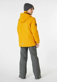 Giacca impermeabile gialla con cappuccio, polsini elasticizzati e dettagli neri, abbinata a pantaloni grigi e un berretto grigio. Sfondo bianco semplice.