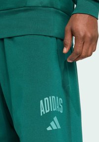 Gröna Adidas sweatpants med en strukturerad yta, som har en vit logotyp på vänster ben. Midjan är elastisk och sitter tajt.