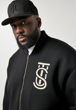 Uomo di colore con barba indossa un cappellino nero dei New York Yankees e una giacca nera con zip con logo bianco ricamato sul petto, su sfondo grigio.