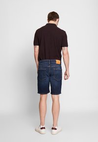 Pantalones cortos de mezclilla azul oscura con corte recto, un ligero desgaste y un parche de cuero marrón en la cinturilla. Zapatillas blancas completan el look.