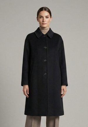 Frau trägt einen schwarz geknöpften, knieumspielenden Mantel über einem beigen Rollkragenpullover und beigen Hosen, steht vor einem schlichten grauen Hintergrund.