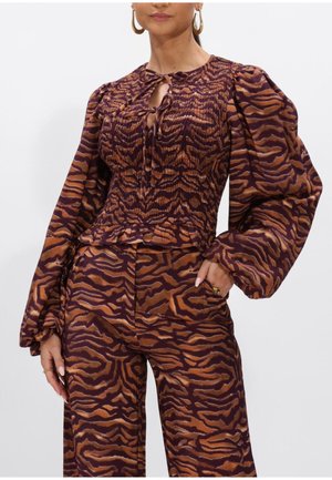 Vrouw draagt een bruin-zwart gestreept topje met tijgerprint en pofmouwen, met bijpassende wijde broek, handen rustend bij de zakken.