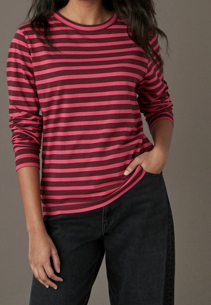 Femme portant une chemise à manches longues rayée rose et rouge foncé et un pantalon noir, avec une main dans la poche, sur un fond uni.