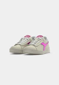 Beige sneakers with a textured surface, pink accent logo, white laces, and a flat rubber sole. Rounded toe design.