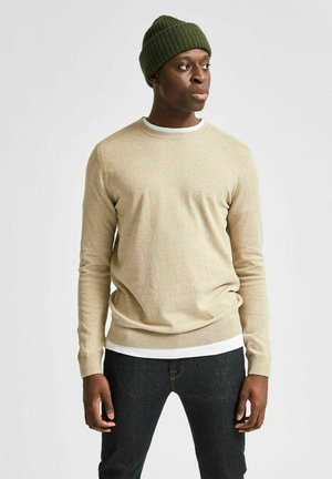 Pull en maille beige avec un col rond et un ourlet côtelé, superposé à une chemise blanche, associé à un jean foncé et un bonnet côtelé vert.